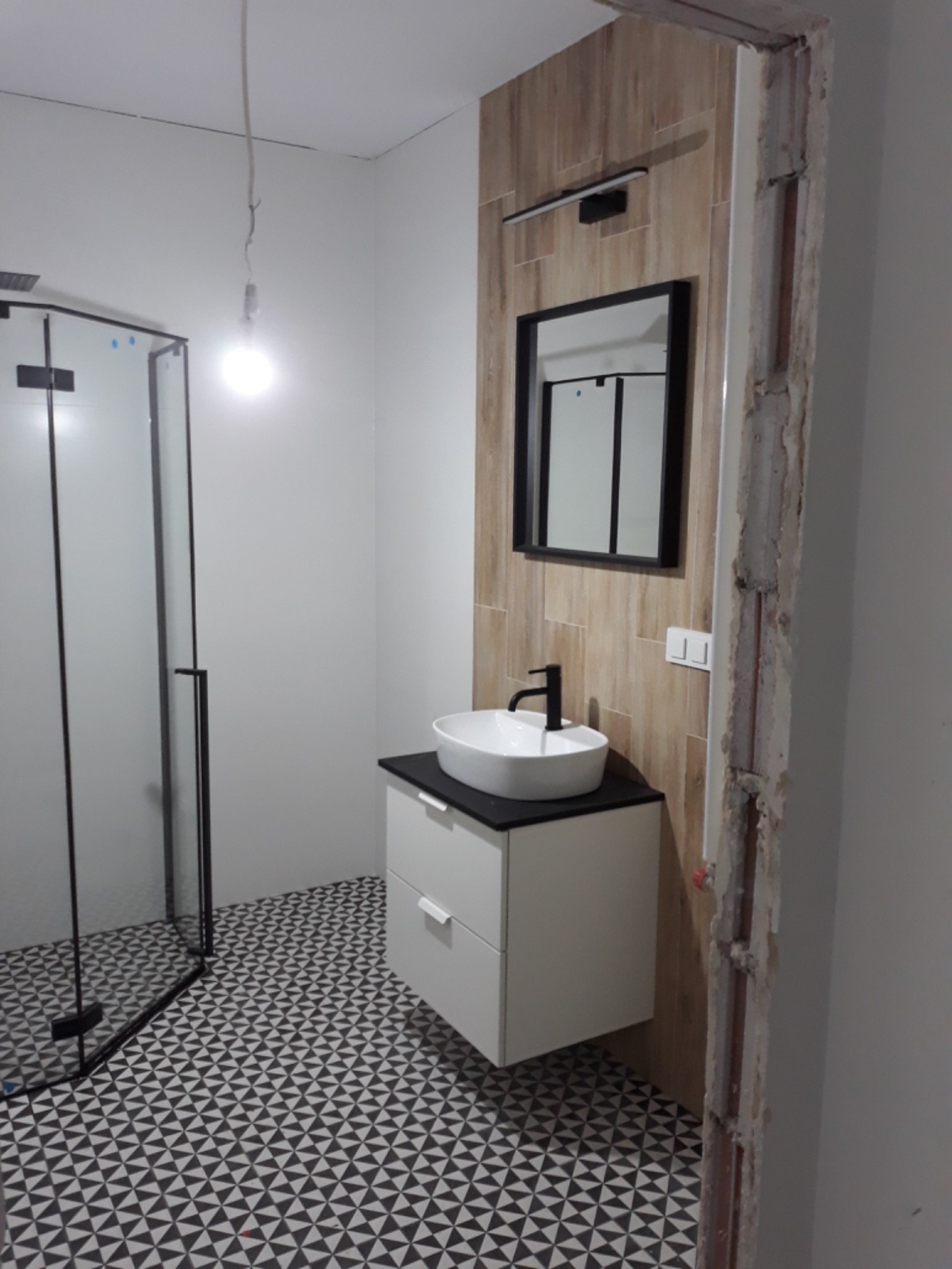 Ukończona łazienka z kabiną prysznicową, umywalką nablatową na szafce z szufladami, lustrem w czarnej ramie i wzorzystą podłogą; widoczne niedokończone wykończenie ościeżnicy.