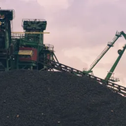 Góra czarnego surowca podawana taśmociągiem do zielonej konstrukcji z podnośnikami na tle pochmurnego nieba.