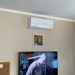 Biały klimatyzator marki AUX zamontowany na ścianie w pokoju, pod nim obrazek z kwiatami, a poniżej włączony telewizor z widocznym mężczyzną ćwiczącym na siłowni.