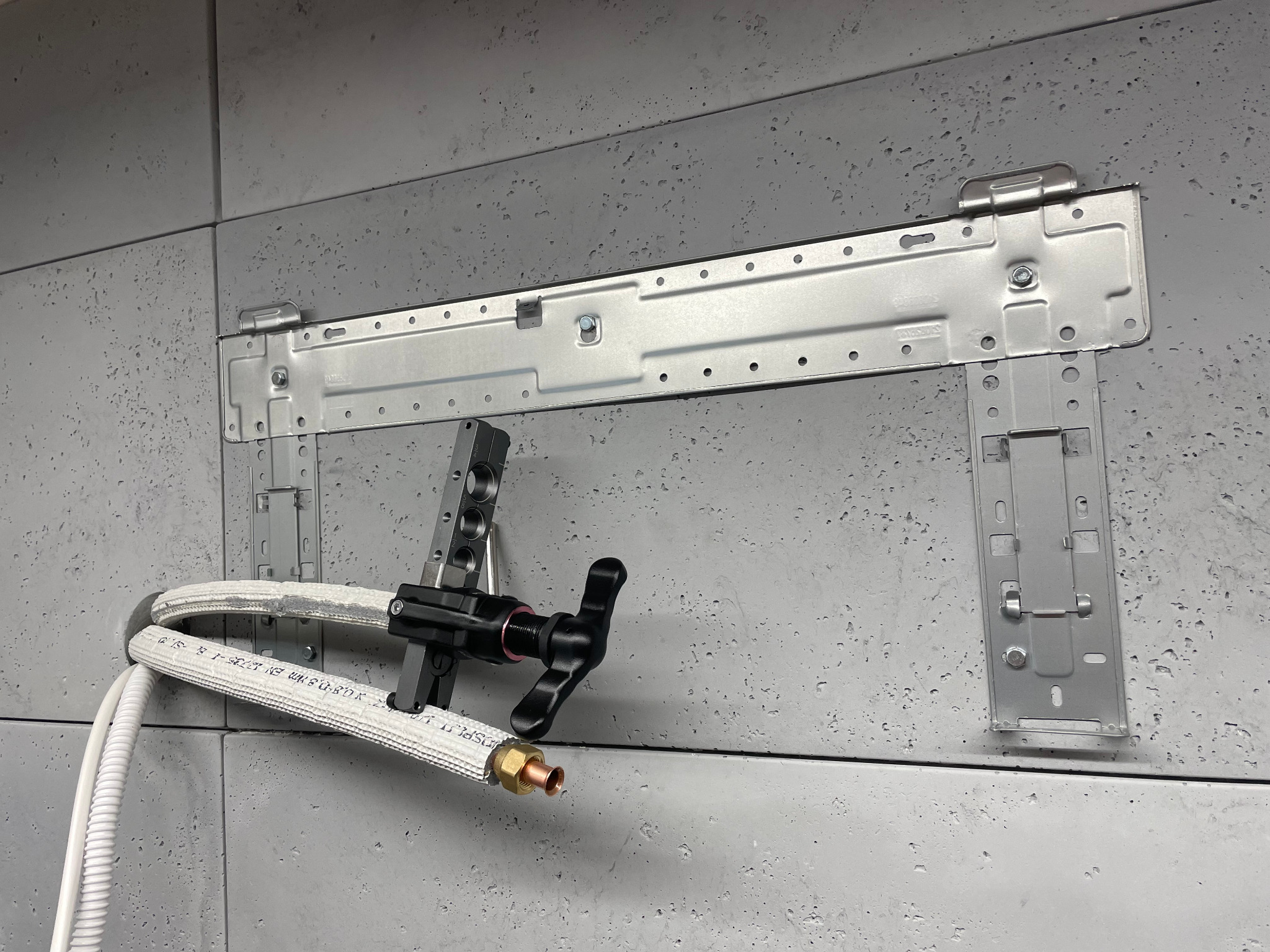 Metalowy wspornik montażowy klimatyzacji przymocowany do szarej ściany, z podłączonymi rurami i zaworem.