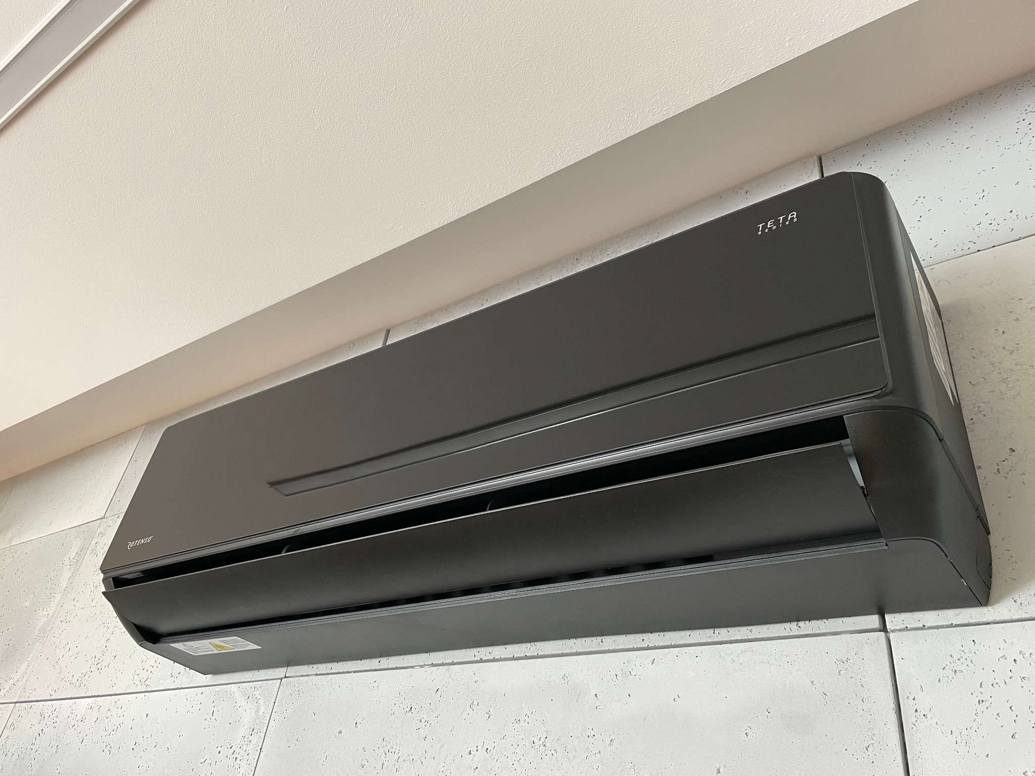 Czarny klimatyzator marki TETA Series zamontowany na ścianie o teksturze betonu, widok z dołu.