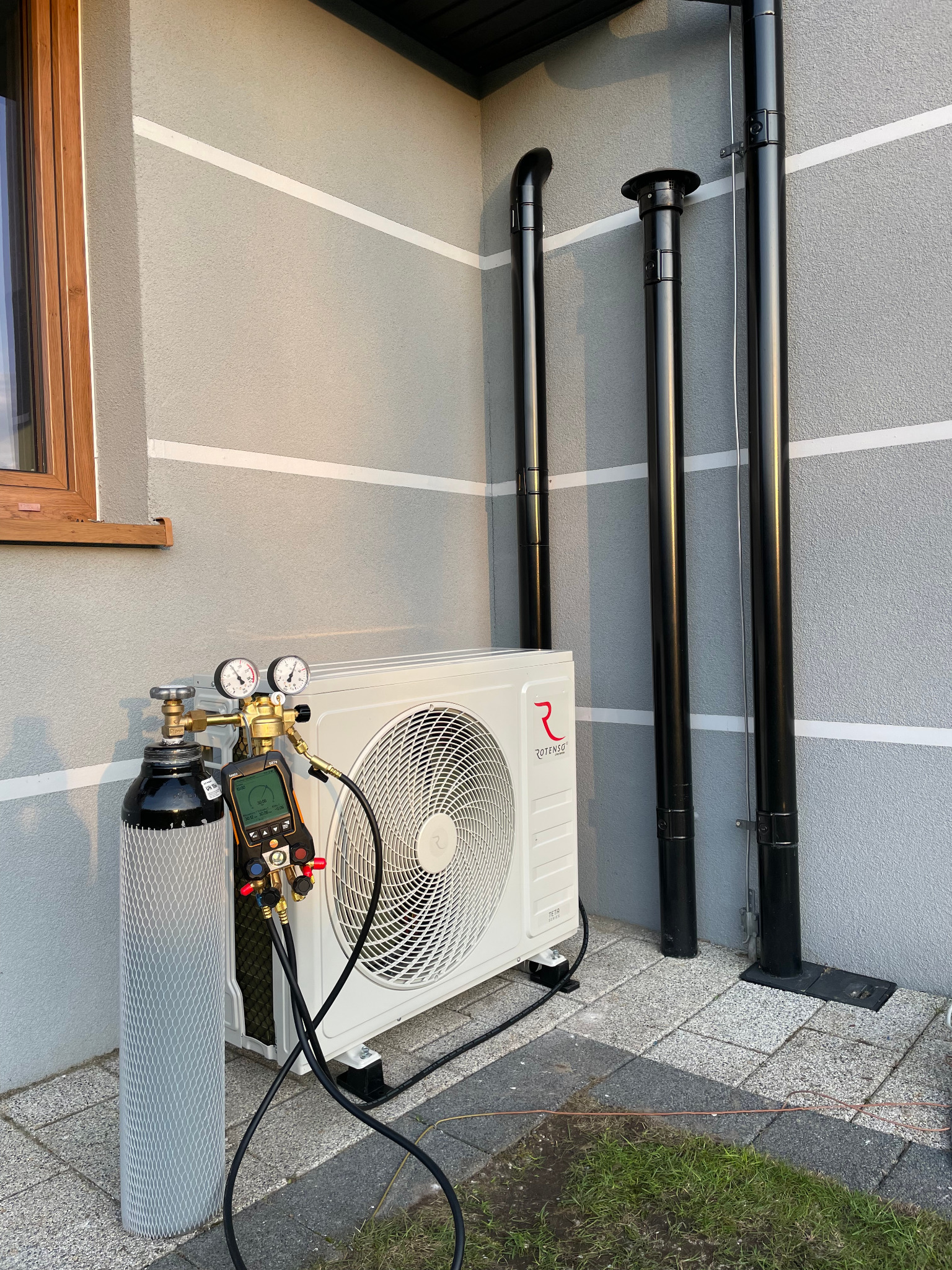 Zewnętrzna jednostka klimatyzacji Rotenso z podłączonym zestawem diagnostycznym i butlą z czynnikiem chłodniczym, ustawiona na kostce brukowej przy ścianie domu z trzema kominami.