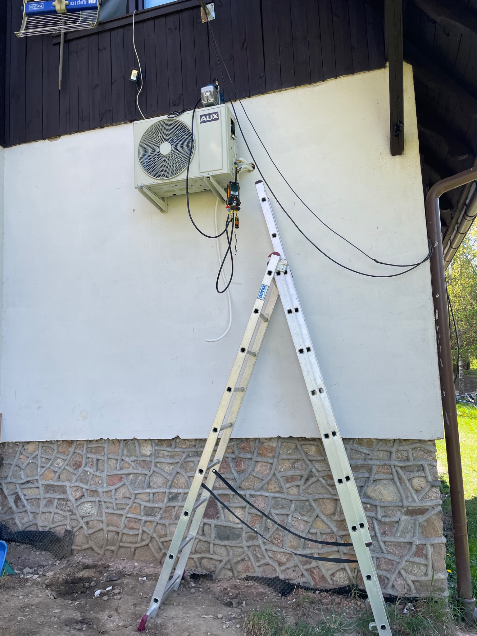 Biała jednostka zewnętrzna klimatyzatora AUX zamontowana na ścianie domu z kamienną podmurówką, widoczna drabina Krause i przewody podłączeniowe.