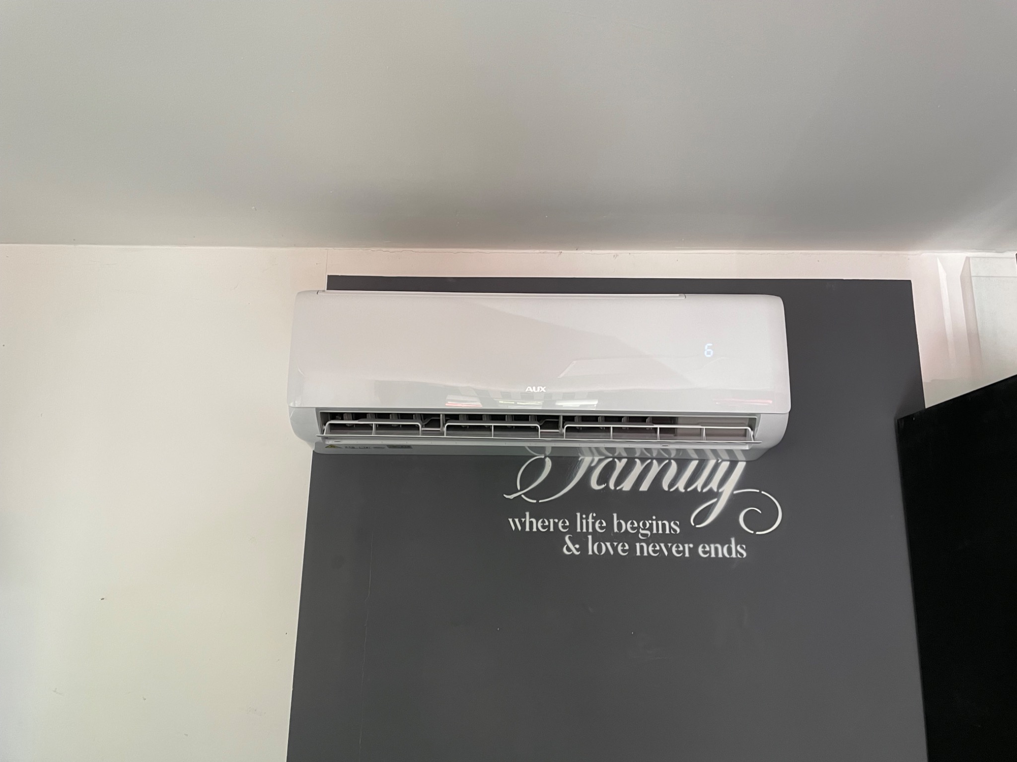 Biały klimatyzator marki AUX zamontowany na szarej ścianie z dekoracyjnym napisem 'Family where life begins & love never ends'.