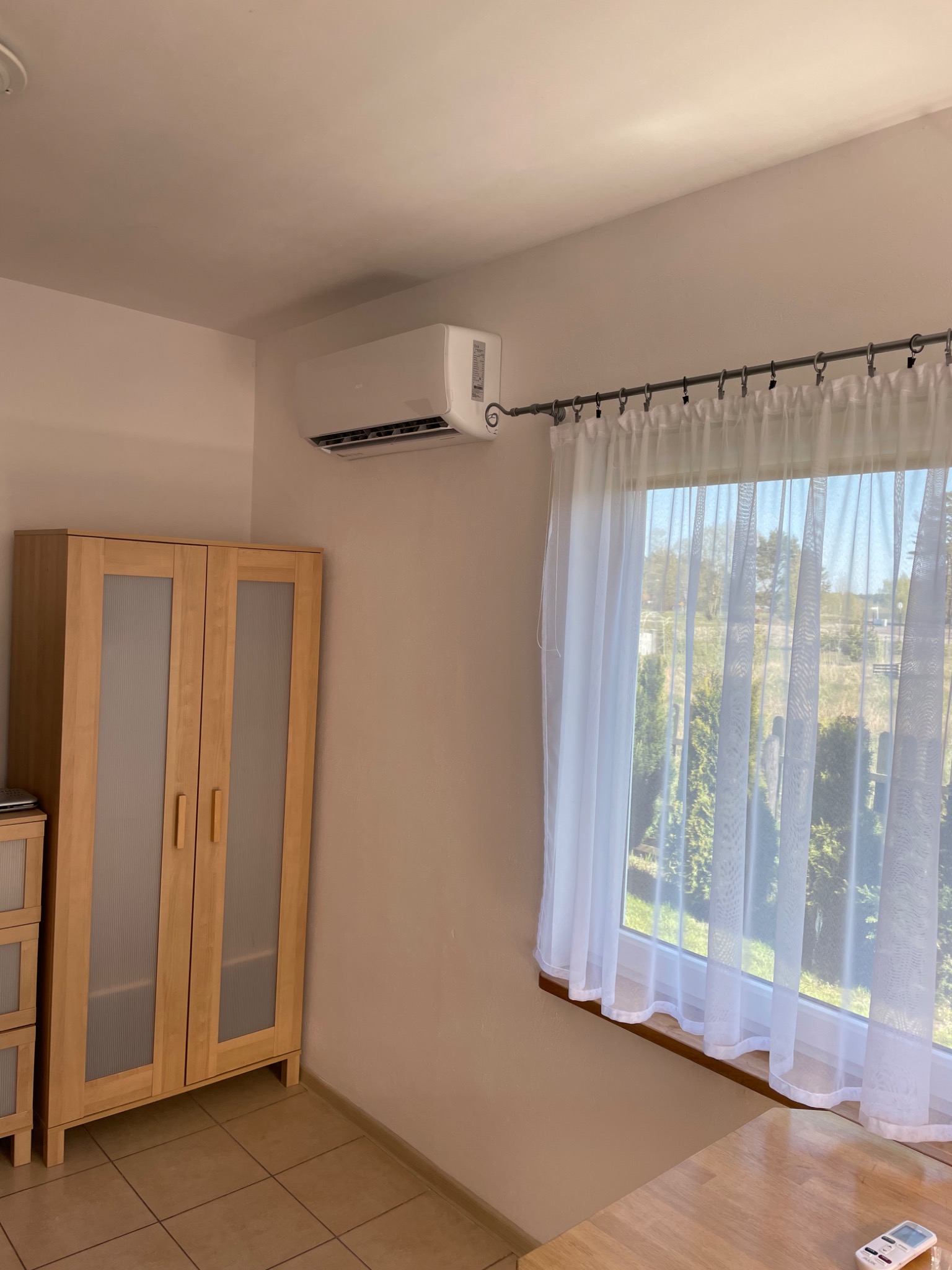 Biały klimatyzator ścienny zamontowany w pokoju z drewnianą szafą i oknem z firankami, widoczny pilot do klimatyzacji na stole.