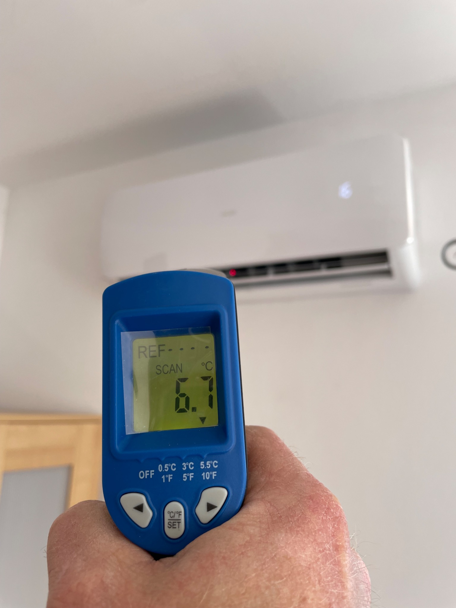 Pomiar temperatury wylotowej klimatyzatora ściennego za pomocą termometru elektronicznego; wyświetlacz wskazuje 6.7 stopni Celsjusza.