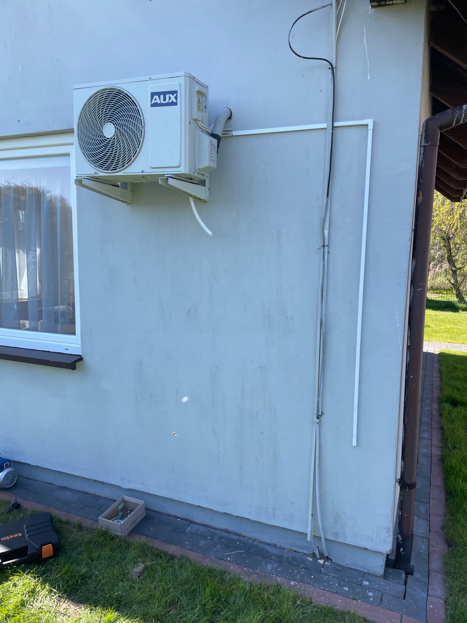 Zamontowany klimatyzator AUX na elewacji budynku jednorodzinnego, widoczne przewody w listwach, skrzynka narzędziowa Testo na trawie.
