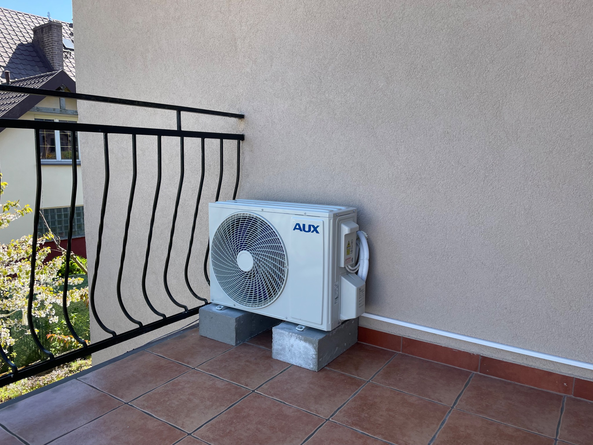 Jednostka zewnętrzna klimatyzacji marki AUX umieszczona na betonowych bloczkach na balkonie z terakotową podłogą i czarną, metalową balustradą. W tle widoczny budynek mieszkalny.
