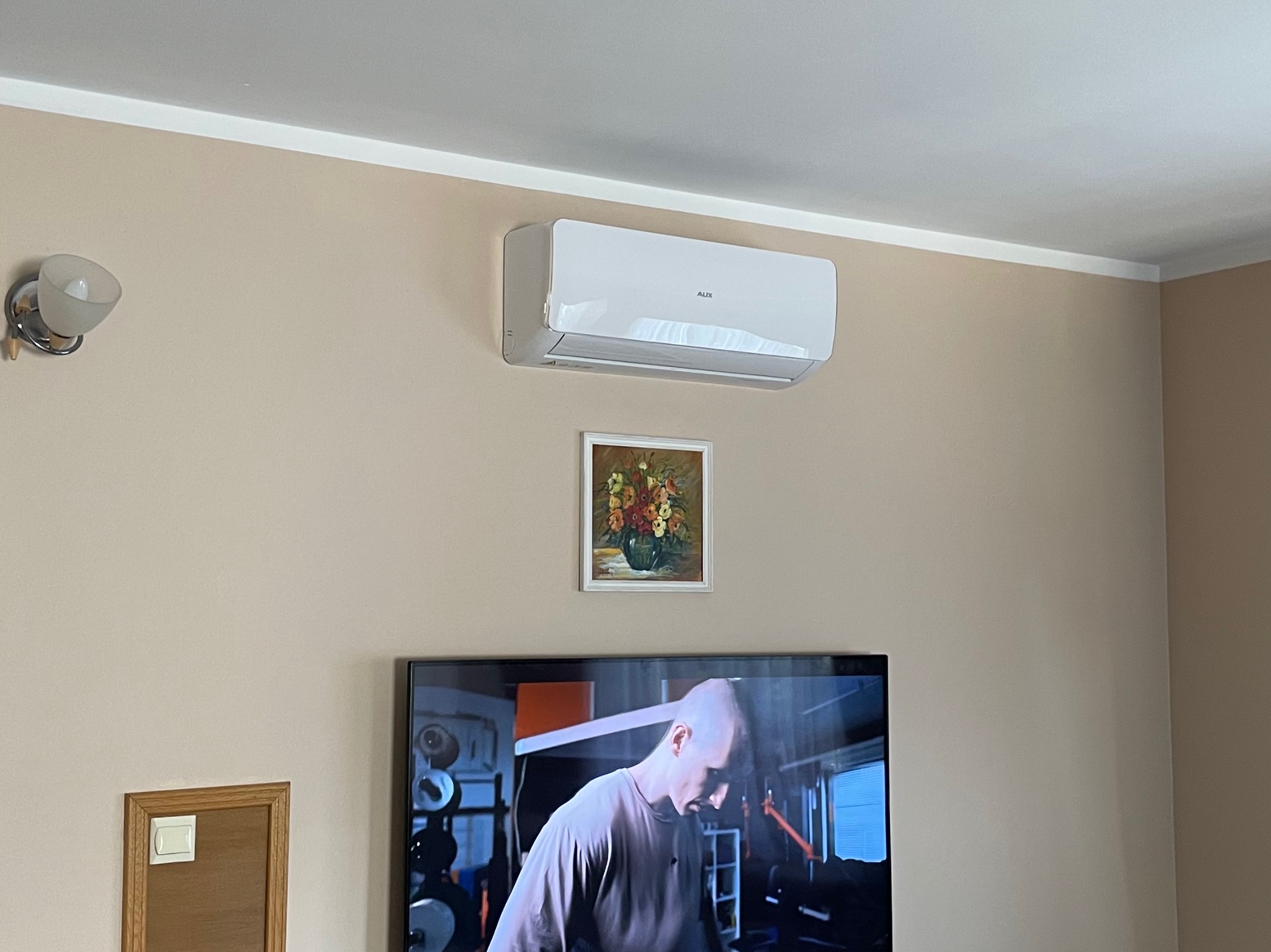 Biały klimatyzator marki AUX zamontowany na ścianie w pokoju, pod nim obrazek z kwiatami, a poniżej włączony telewizor z widocznym mężczyzną ćwiczącym na siłowni.