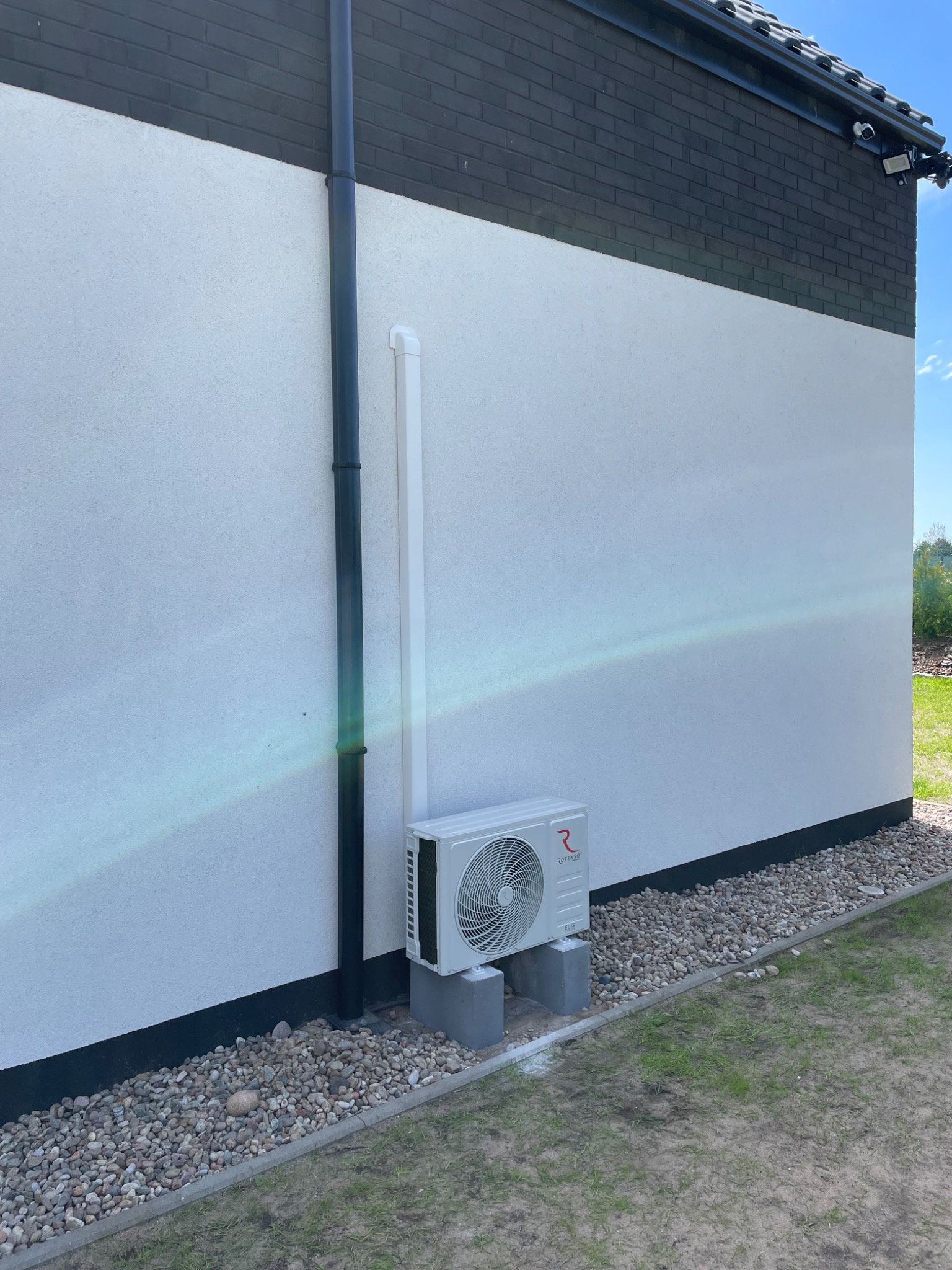Zewnętrzna jednostka klimatyzacji Rotenso ELIS zamontowana na betonowych bloczkach przy ścianie domu z orynnowaniem i kamerą monitoringu.