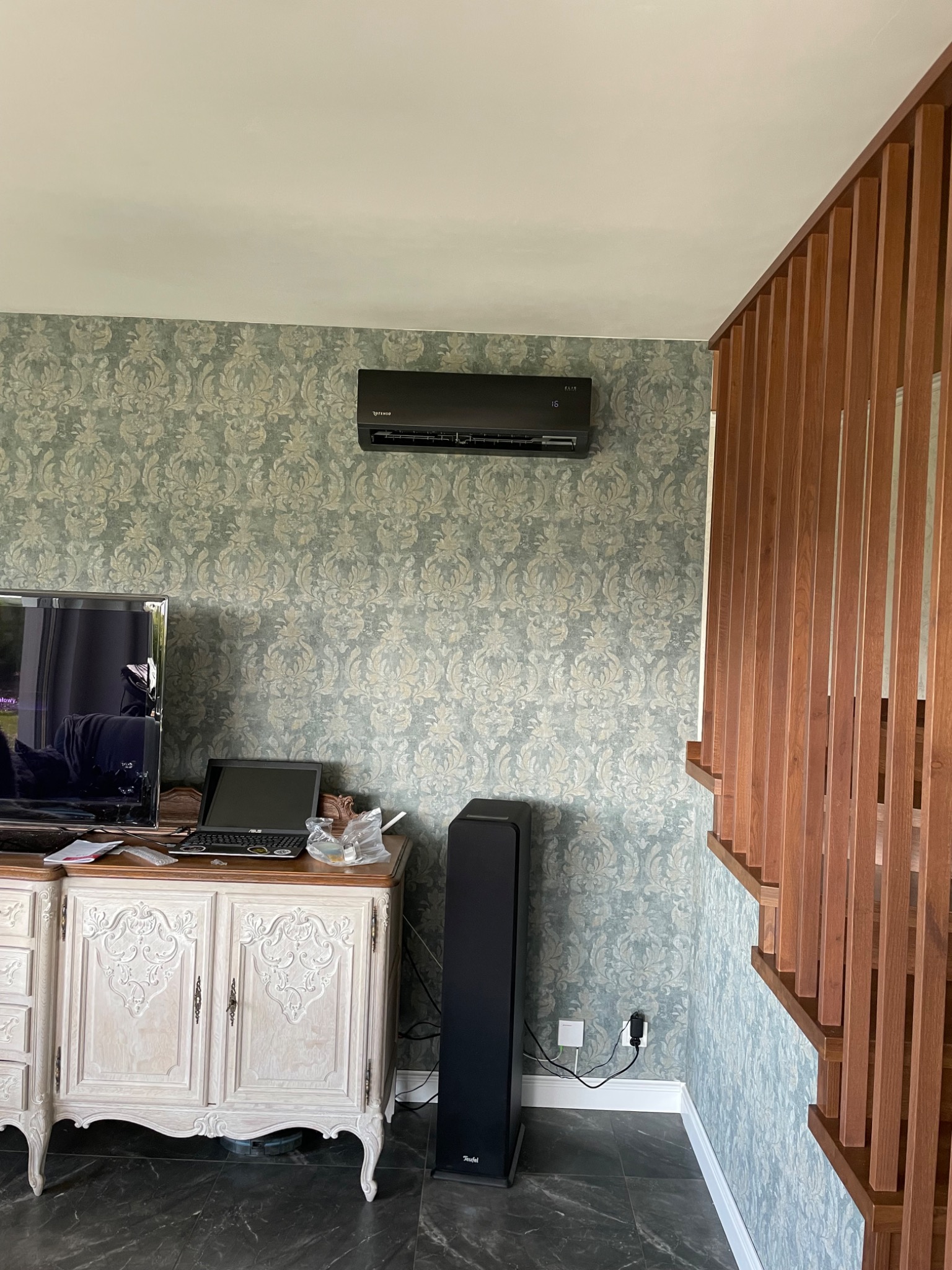 Czarny klimatyzator ścienny zamontowany na ścianie z wzorzystą tapetą w salonie, widoczny fragment drewnianej balustrady schodów, szafka RTV z laptopem i głośnik.