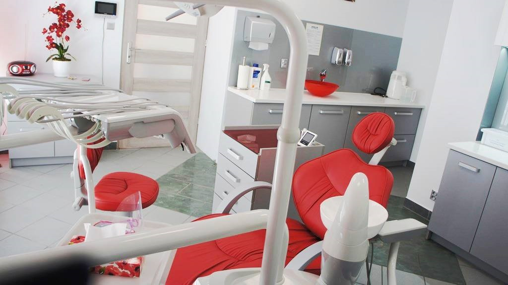 Czerwony fotel dentystyczny w nowoczesnym gabinecie, widoczne narzędzia i akcesoria, na blacie czerwona misa i dozowniki, w tle roślina doniczkowa.