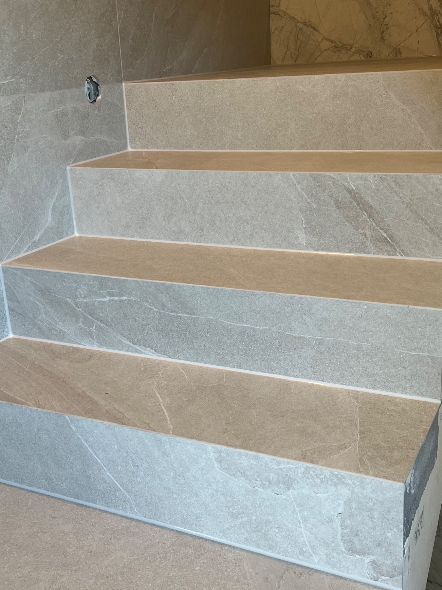 Wykończone schody w łazience z płytek imitujących marmur, z widocznym otworem na instalację elektryczną w ścianie i niedokończonym wykończeniem na dole.