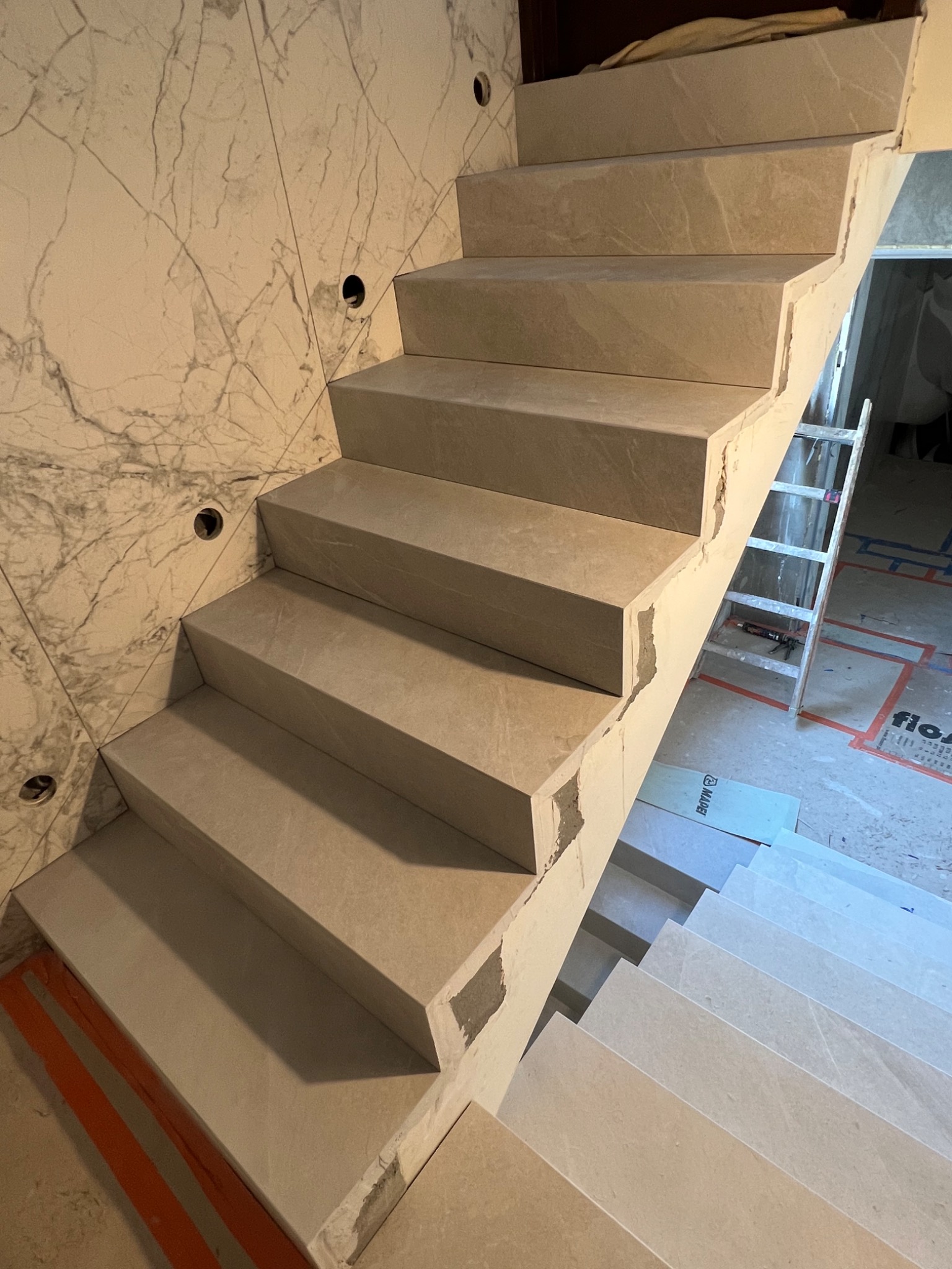 Montaż płytek imitujących kamień na schodach wewnętrznych, widoczne niedociągnięcia i resztki kleju, marmurowa okładzina ściany z otworami na oświetlenie.