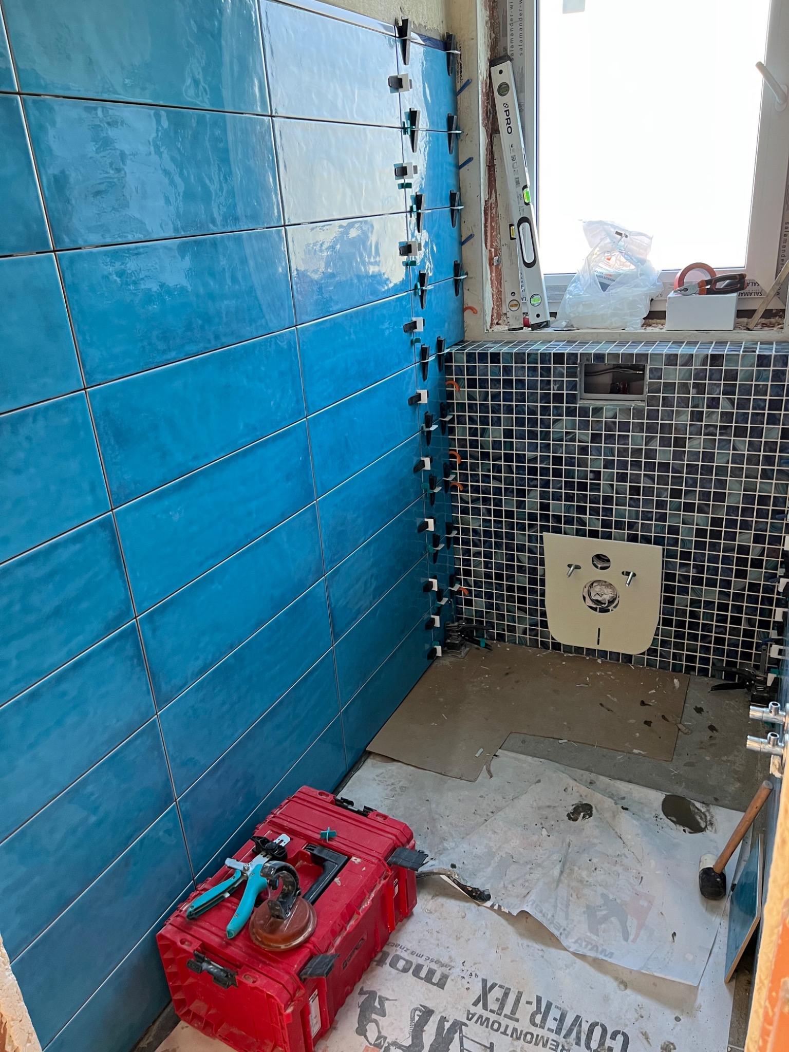 Ściana łazienki w trakcie układania niebieskich płytek, widoczne krzyżyki dystansowe, poziomica przy oknie, oraz czerwona skrzynka z narzędziami glazurniczymi.