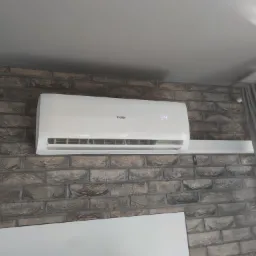 Biały klimatyzator Haier zamontowany na ścianie z cegły, z wyświetlaczem pokazującym temperaturę 24 stopnie Celsjusza, obok zasłony.