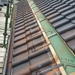 Widok z góry na fragment dachu w trakcie budowy, z czarną dachówką, elementami montażowymi, deskami i zieloną membraną, widoczne rusztowanie po lewej stronie.