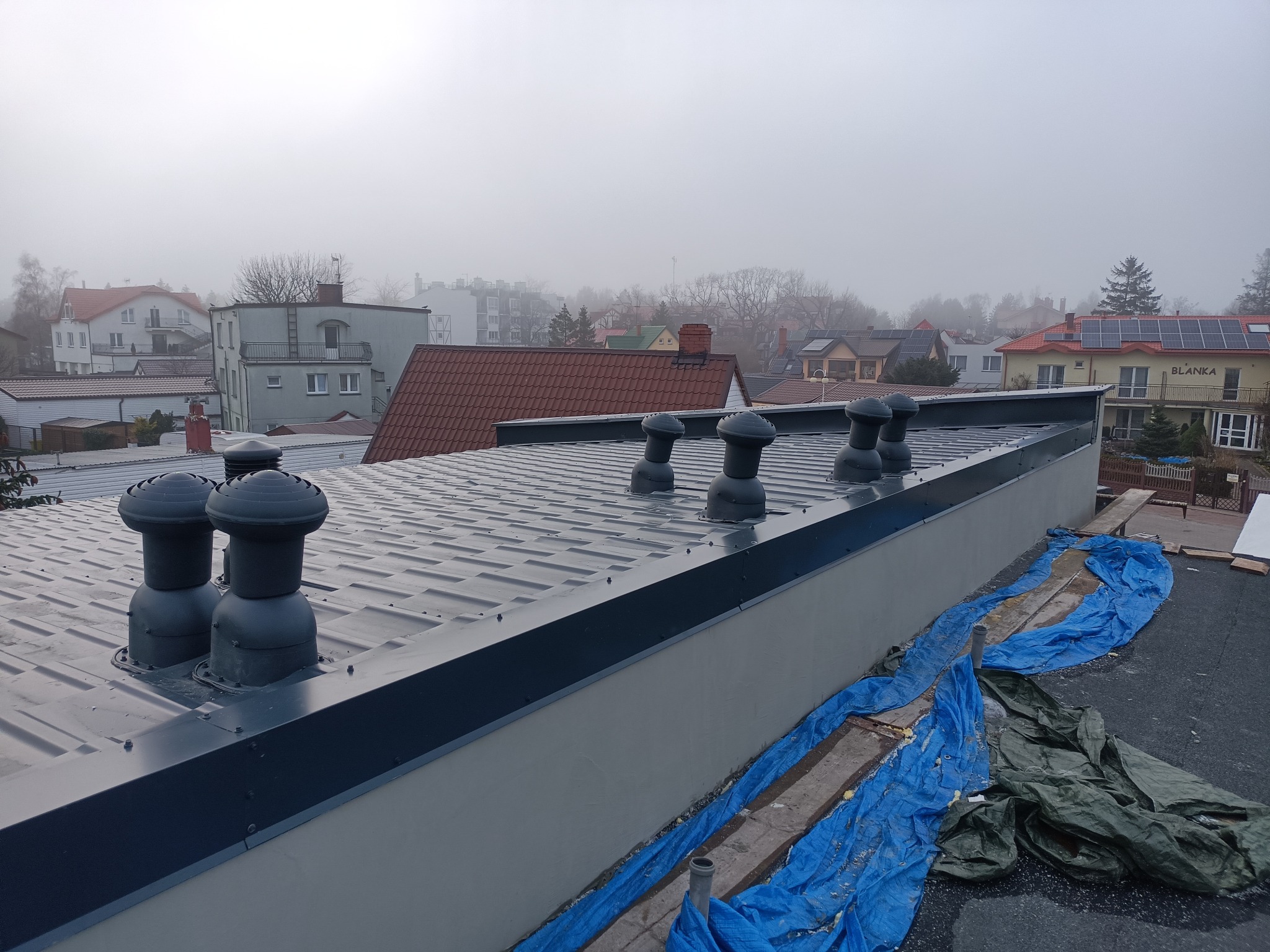 Szary dach z blachy trapezowej z sześcioma kominkami wentylacyjnymi na tle zabudowy miejskiej spowitej mgłą, zabezpieczony plandekami. Widoczne elementy obróbki blacharskiej.
