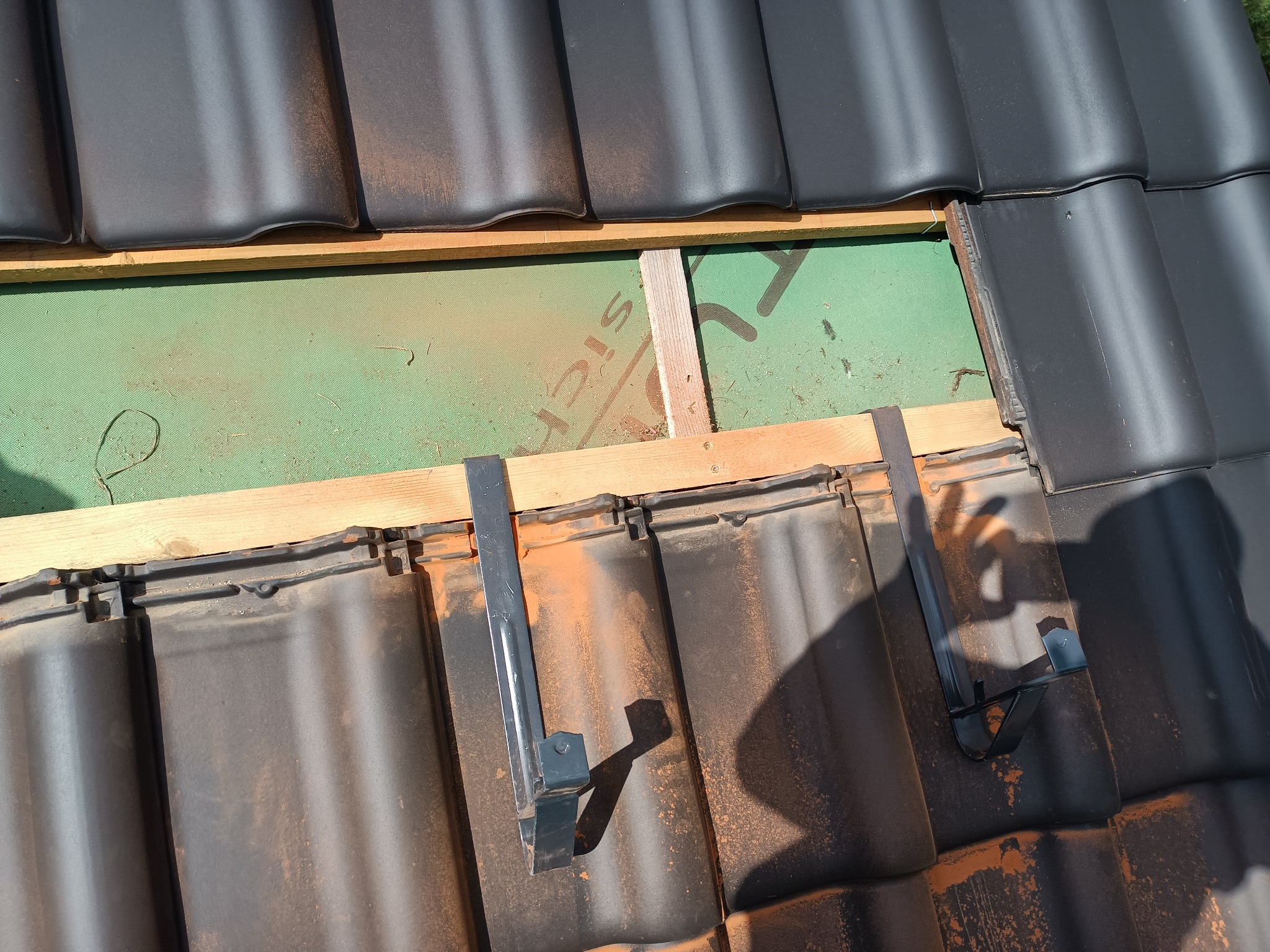 Montaż czarnej dachówki ceramicznej na drewnianej konstrukcji z widoczną membraną dachową i metalowymi uchwytami, cień osoby w prawym rogu.