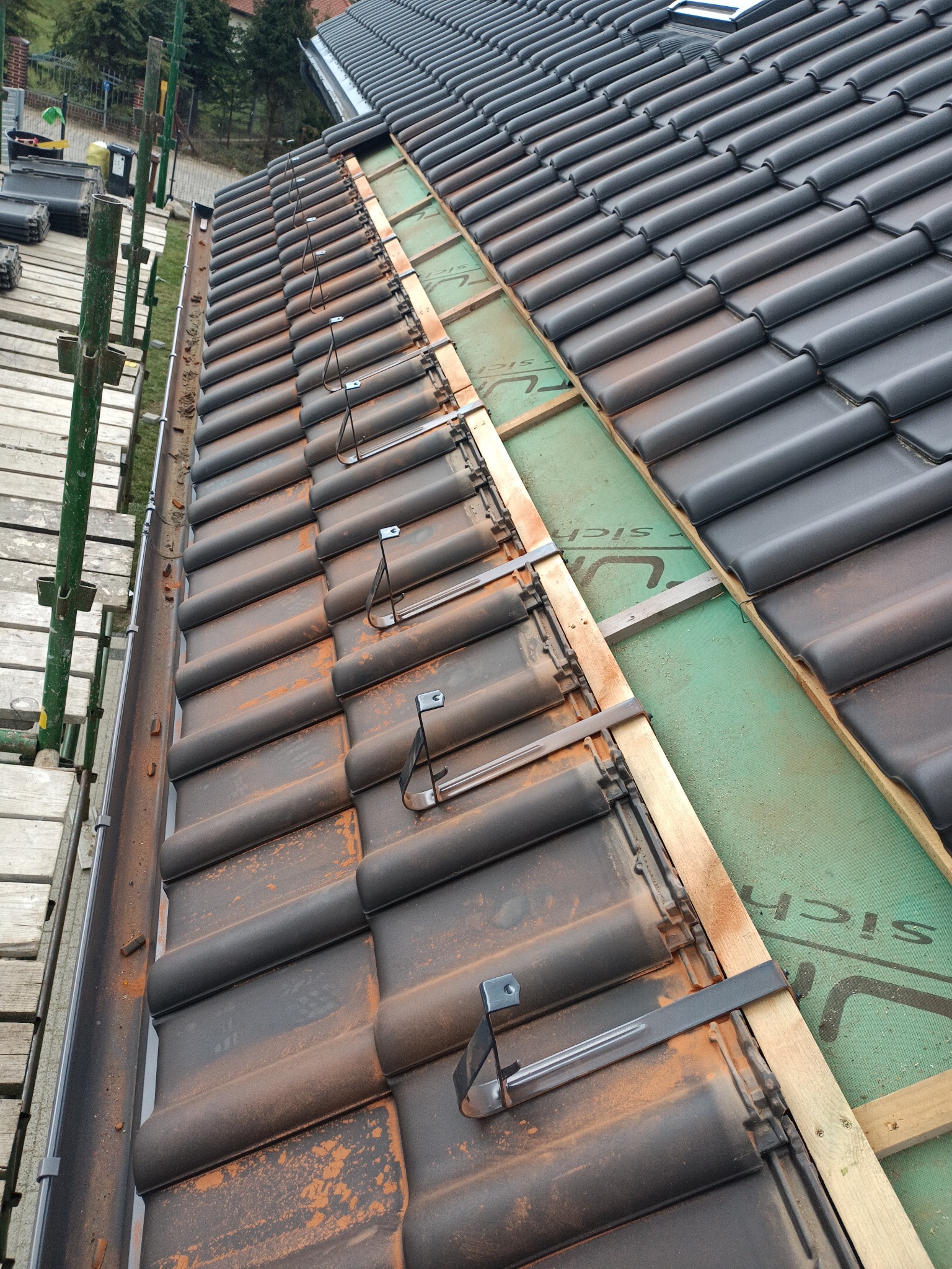 Widok z góry na fragment dachu w trakcie budowy, z czarną dachówką, elementami montażowymi, deskami i zieloną membraną, widoczne rusztowanie po lewej stronie.