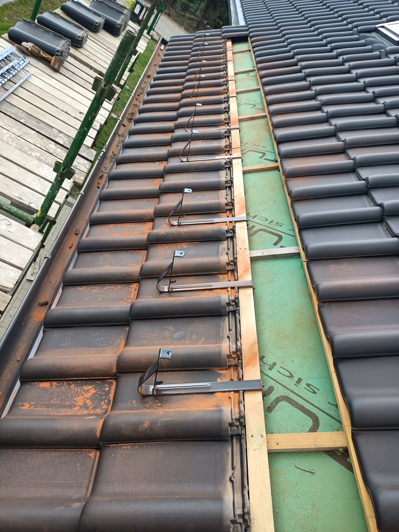 Montaż czarnej dachówki z metalowymi uchwytami na łatach w Niepoczołowicach, widoczne rusztowanie i zielona membrana dachowa.