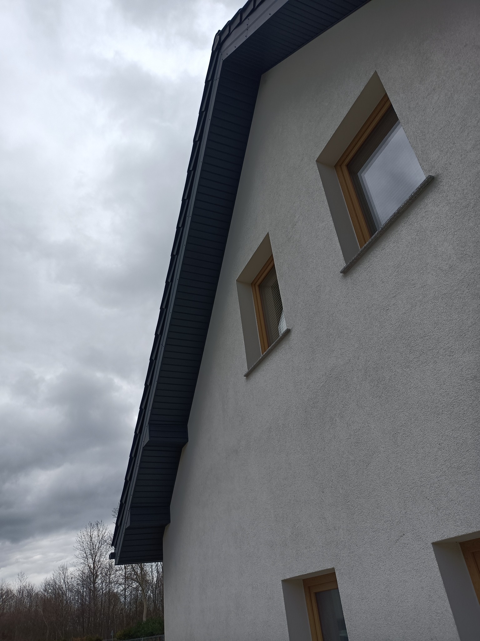 Fragment elewacji budynku z oknami i wystającym okapem dachu na tle pochmurnego nieba.
