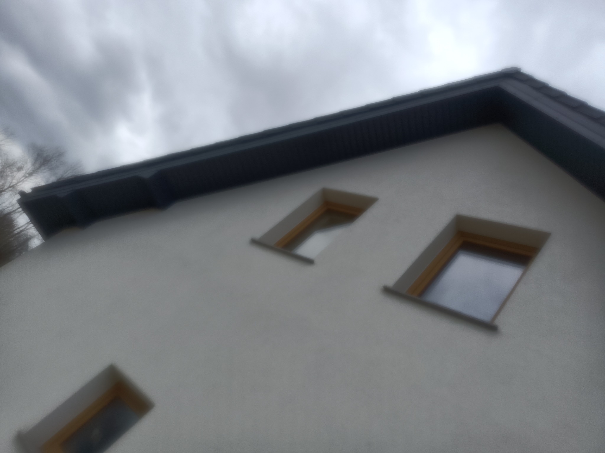 Fragment elewacji budynku z trzema oknami dachowymi, widok z dołu na szary dach i pochmurne niebo.