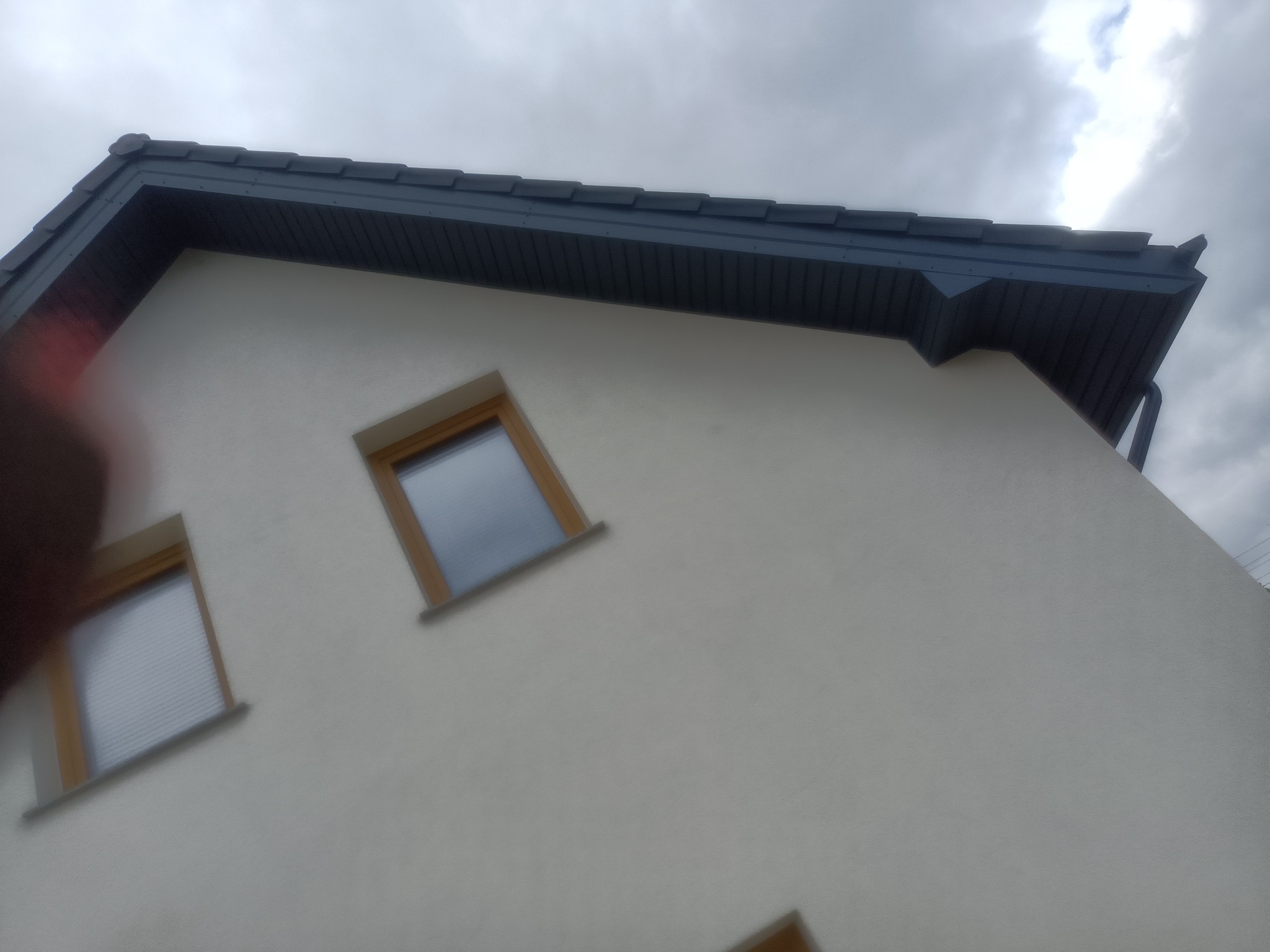 Fragment elewacji budynku z dwoma oknami z drewnianymi ramami i szarym dachem z widocznymi rynnami, sfotografowany pod kątem z dołu w pochmurny dzień.