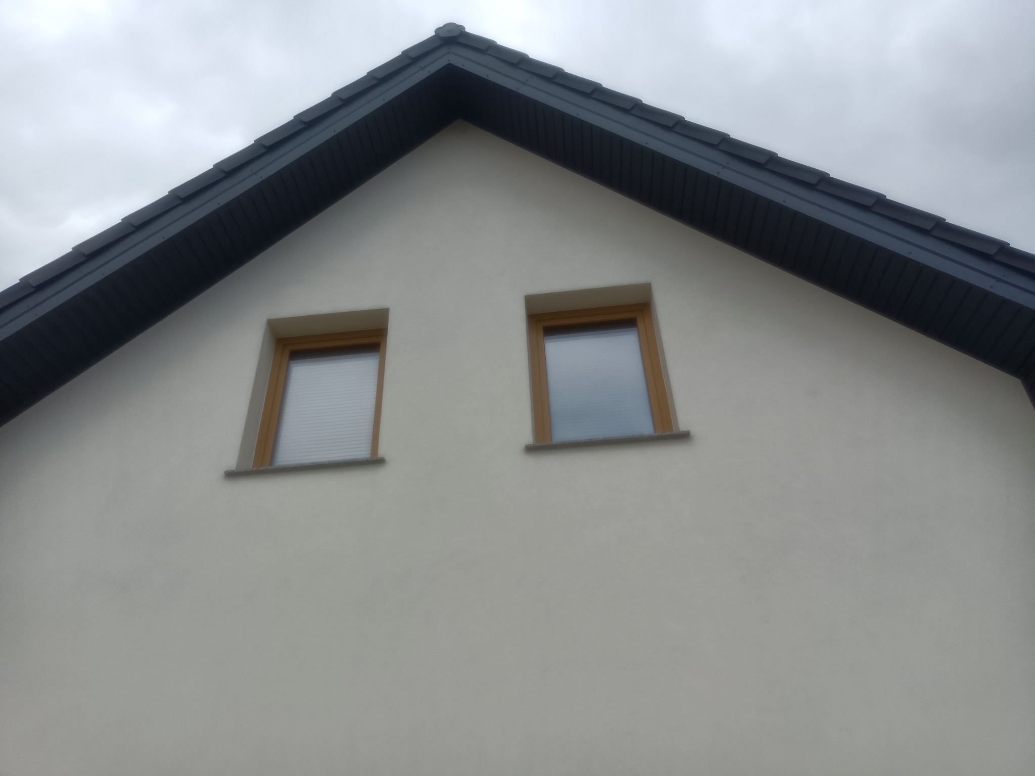 Fragment szczytu domu z nowym, ciemnoszarym dachem z dachówki ceramicznej i dwoma oknami poddaszowymi z drewnianą ramą, widziany z dołu na tle pochmurnego nieba.