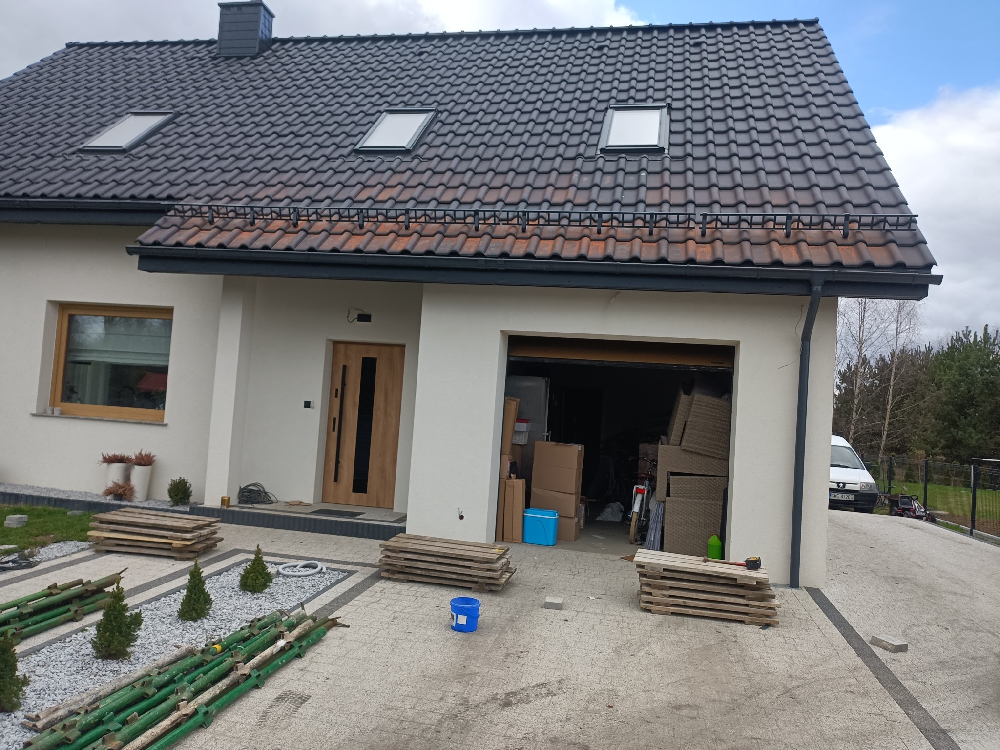 Dom jednorodzinny z ciemną dachówką ceramiczną, trzema oknami dachowymi i orynnowaniem. Na podjeździe leżą elementy rusztowania i palety. Otwarty garaż wypełniony meblami ogrodowymi i kartonami.