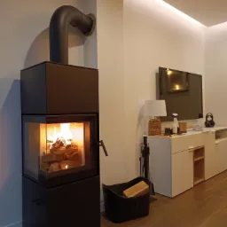 Nowoczesny, czarny kominek wolnostojący z palącym się drewnem, z widocznym przewodem kominowym i koszem na drewno w salonie z białymi meblami.