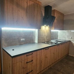 Kamil Maliszewski - Nowoczesna kuchnia z drewnianymi frontami szafek, czarnym blatem i okapem, oświetlona listwą LED pod szafkami wiszącymi, z płytkami imitującymi beton na ścianie.