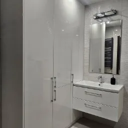 Biała, lakierowana szafa łazienkowa na wymiar z umywalką nablatową i dwoma szufladami, uzupełniona o lustro z oświetleniem i grzejnik drabinkowy w tle.