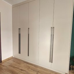 Kamil Maliszewski - Jasna zabudowa szafy wnękowej z długimi, czarnymi uchwytami, dopasowana do wysokości pomieszczenia, na tle drewnianej podłogi.