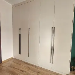 Jasna zabudowa szafy wnękowej z długimi, czarnymi uchwytami, dopasowana do wysokości pomieszczenia, na tle drewnianej podłogi.