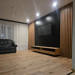 Nowoczesny salon z czarną ścianą telewizyjną, drewnianymi lamelami, podwieszaną szafką RTV i parkietem, oświetlony punktowymi lampami sufitowymi, z czarną skórzaną sofą w tle.