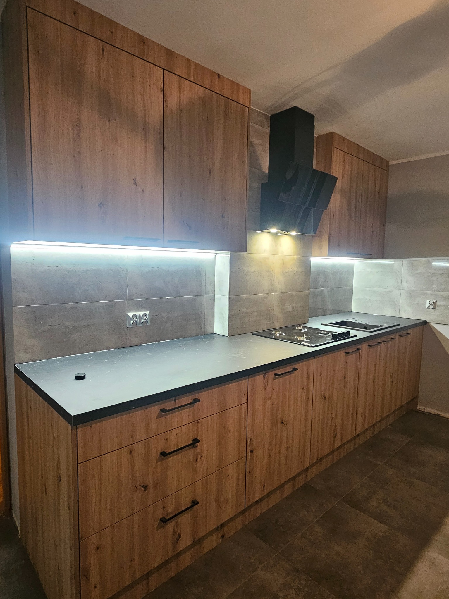 Nowoczesna kuchnia z drewnianymi frontami szafek, czarnym blatem i okapem, oświetlona listwą LED pod szafkami wiszącymi, z płytkami imitującymi beton na ścianie.