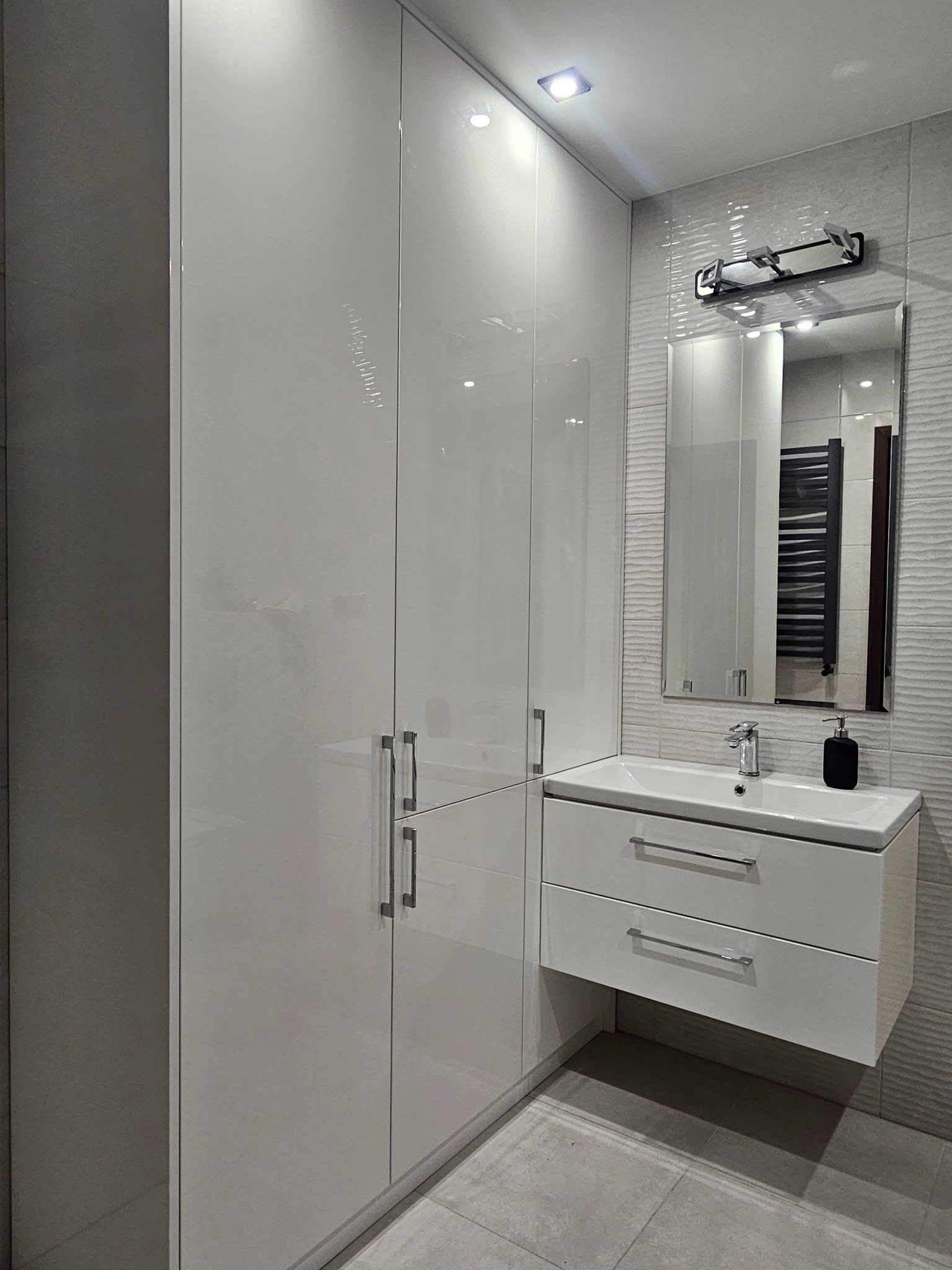 Biała, lakierowana szafa łazienkowa na wymiar z umywalką nablatową i dwoma szufladami, uzupełniona o lustro z oświetleniem i grzejnik drabinkowy w tle.