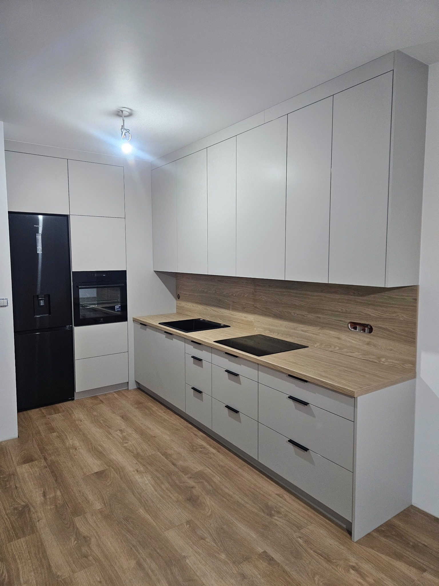 Nowoczesna kuchnia w odcieniach szarości z drewnianym blatem i czarnymi akcentami, widok na zabudowę z lodówką i piekarnikiem, oraz płytą indukcyjną i zlewozmywakiem.