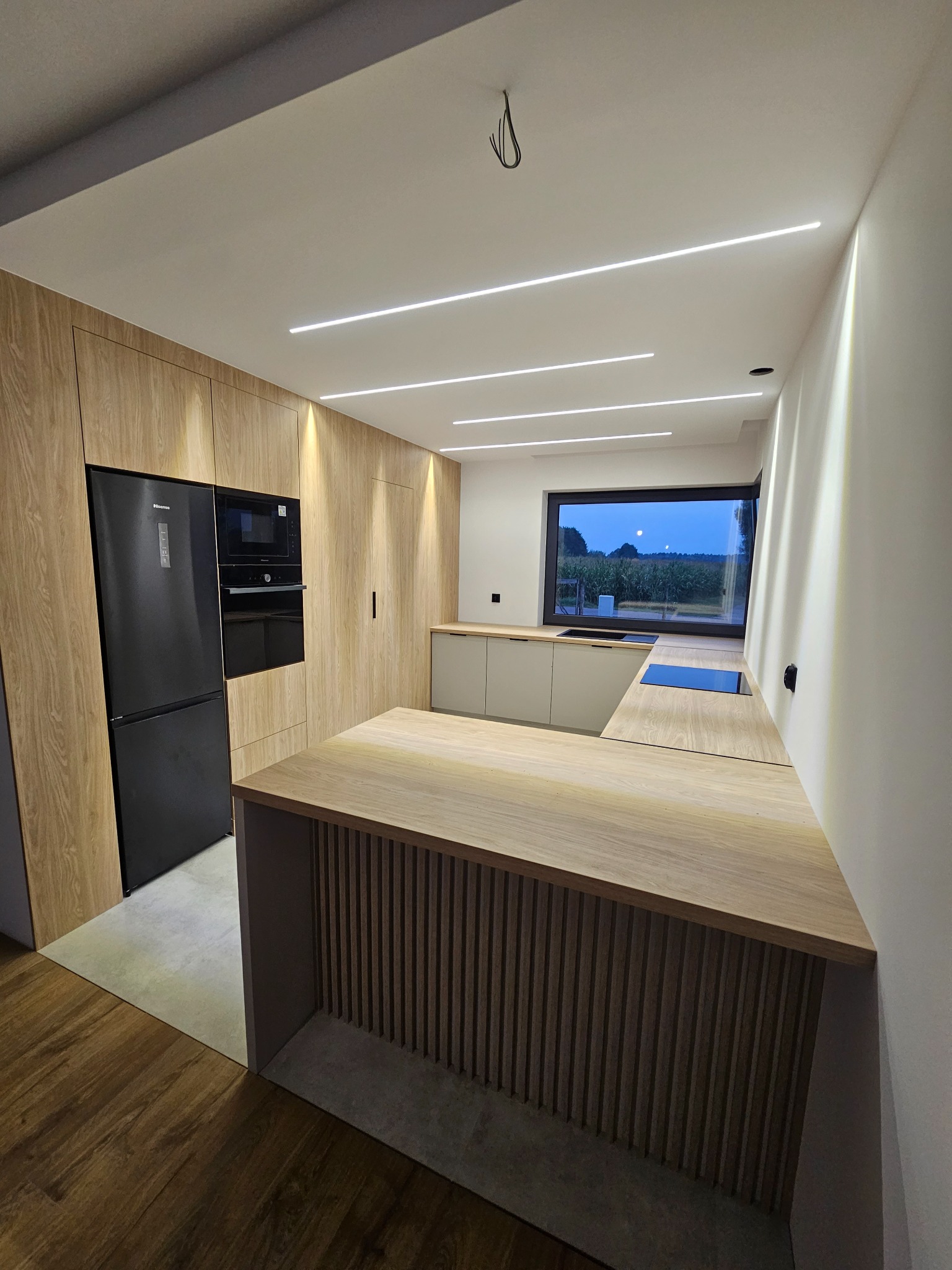 Nowoczesna kuchnia z drewnianymi frontami, czarnym sprzętem AGD i oświetleniem LED, widok na krajobraz za oknem.