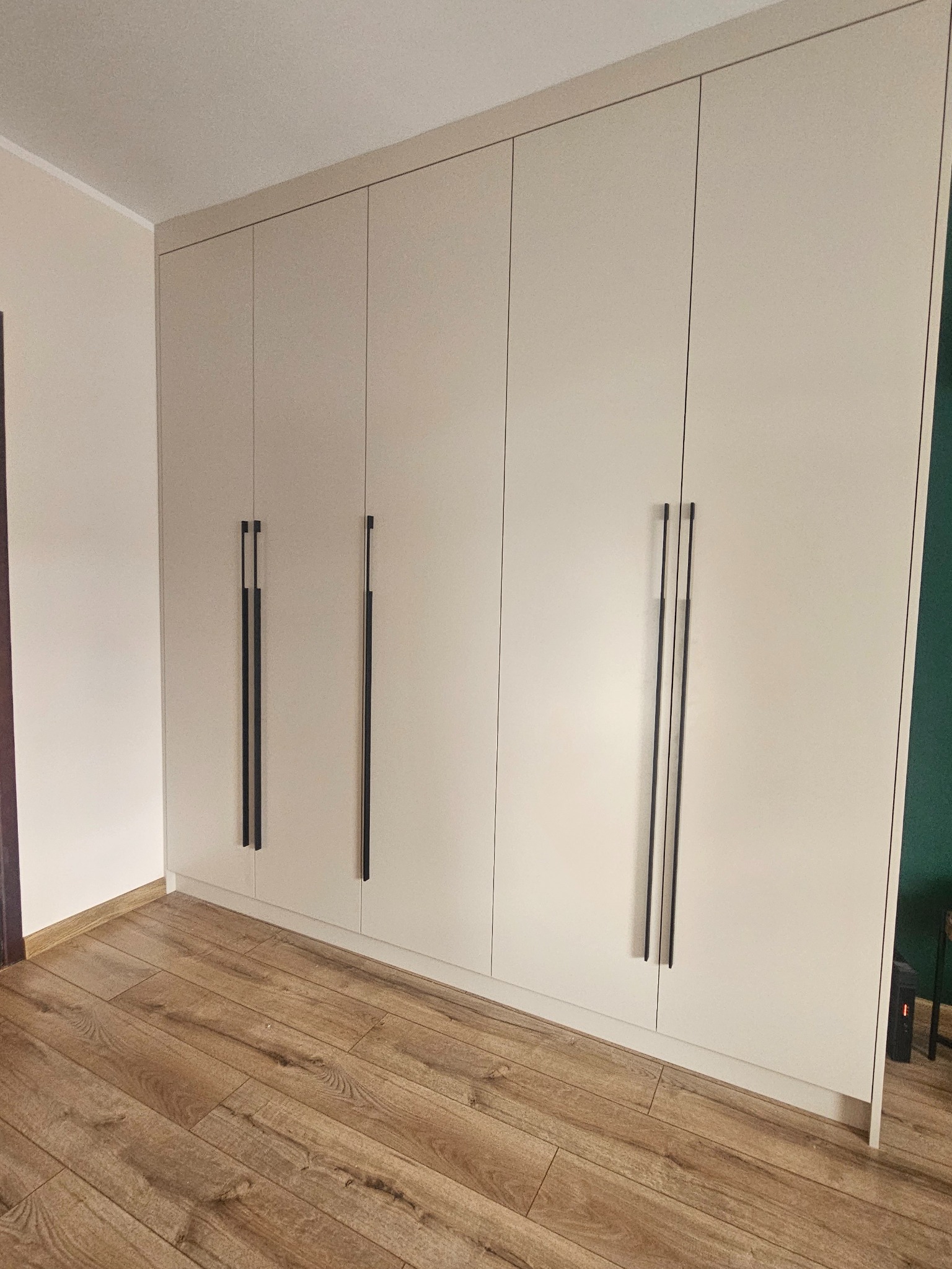 Jasna zabudowa szafy wnękowej z długimi, czarnymi uchwytami, dopasowana do wysokości pomieszczenia, na tle drewnianej podłogi.