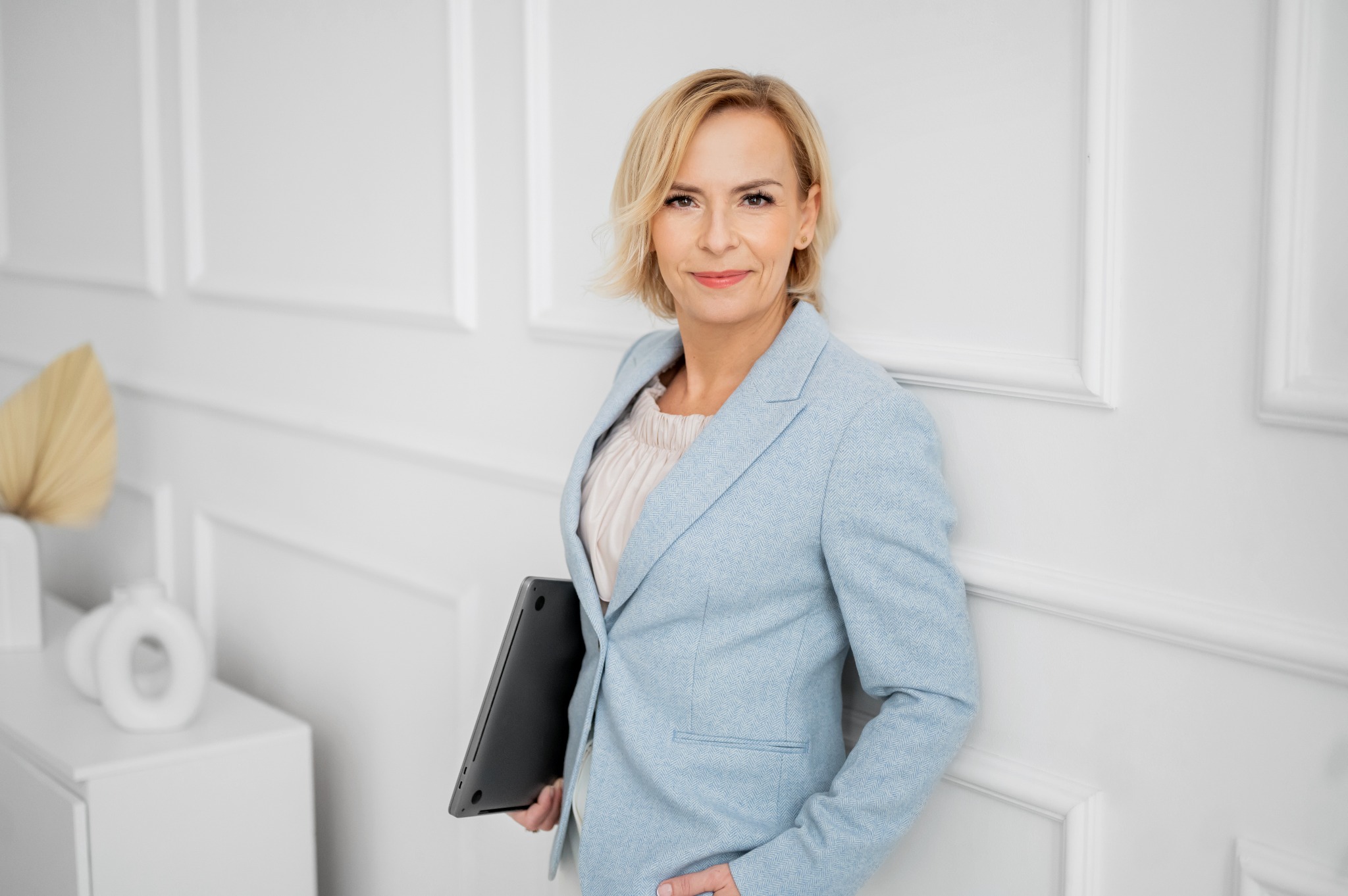 Uśmiechnięta blondynka w błękitnej marynarce, oparta o białą ścianę z dekoracyjnymi panelami, trzyma zamkniętego laptopa pod pachą.