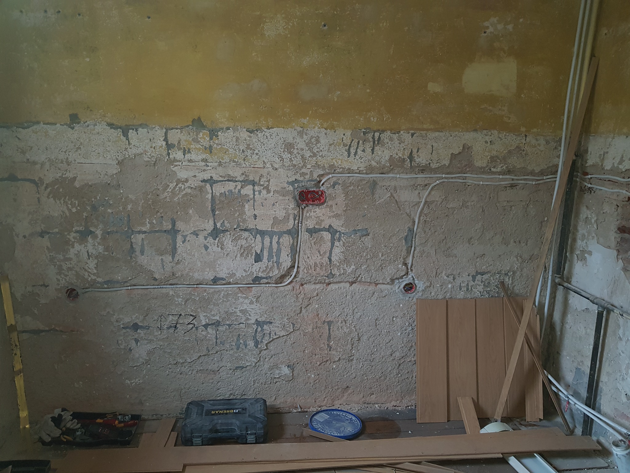 Ściana w trakcie remontu z widocznymi przewodami elektrycznymi w peszlach, puszkami instalacyjnymi, skrzynka narzędziowa i panele podłogowe w surowym wnętrzu.