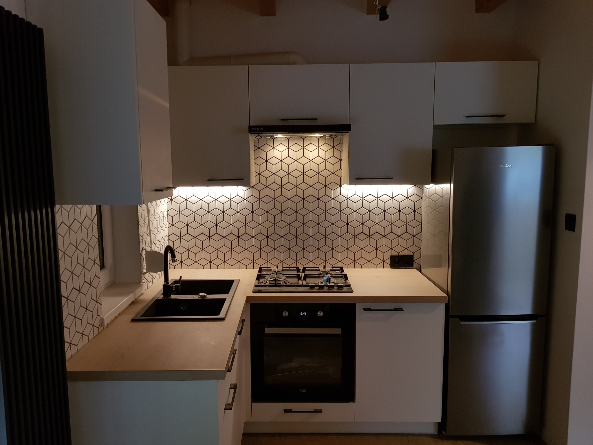 Nowoczesna kuchnia z białymi szafkami, drewnianym blatem, czarnym zlewem i płytą gazową z piekarnikiem, oświetlona lampkami podszafkowymi i lampą nad okapem, z geometryczną mozaiką na ścianie.