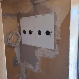 Dwa białe panele elektryczne z okrągłymi otworami i kluczami w zamkach, zamontowane na surowej, otynkowanej ścianie w odcieniach beżu, z widocznymi śladami po pracach wykończeniowych i zwisającym...