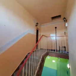 Klatka schodowa w bloku mieszkalnym, widoczne lampy ścienne, pomalowane ściany z widocznymi śladami po remoncie, czerwona poręcz, schody w dół z zabezpieczeniem i zielona dolna część klatki schodowej.