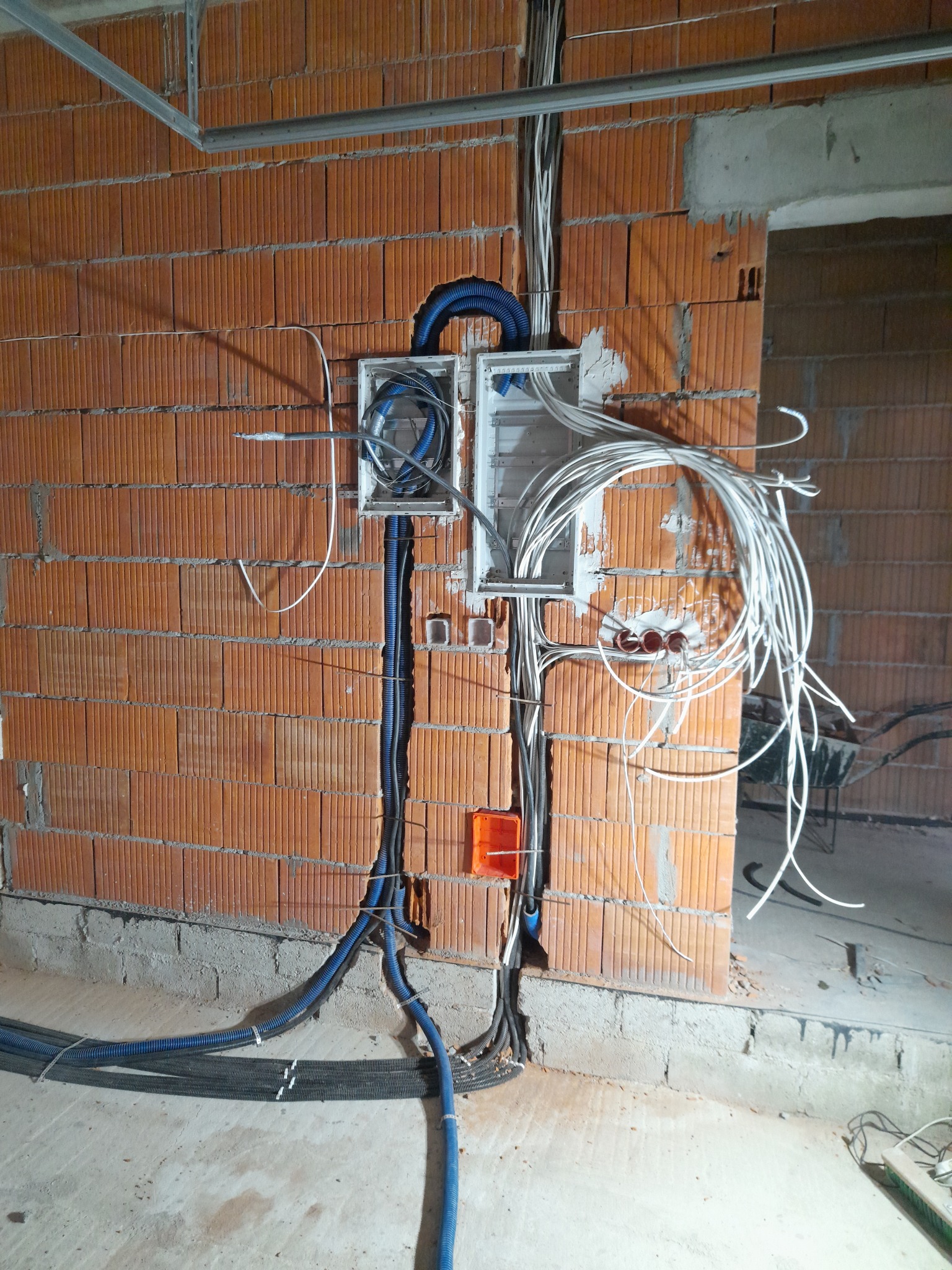 Rozdzielnica elektryczna w trakcie montażu w surowym wnętrzu, kable w peszlach prowadzone wzdłuż ceglanej ściany, widoczne elementy konstrukcji sufitu podwieszanego i taczka w tle.