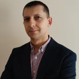 Portret mężczyzny w ciemnej marynarce i koszuli w czerwoną i niebieską kratę, z neutralnym wyrazem twarzy na jasnym tle.