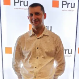 Portret uśmiechniętego mężczyzny w koszuli na tle z logo firmy ubezpieczeniowej Pru, część M&G plc.