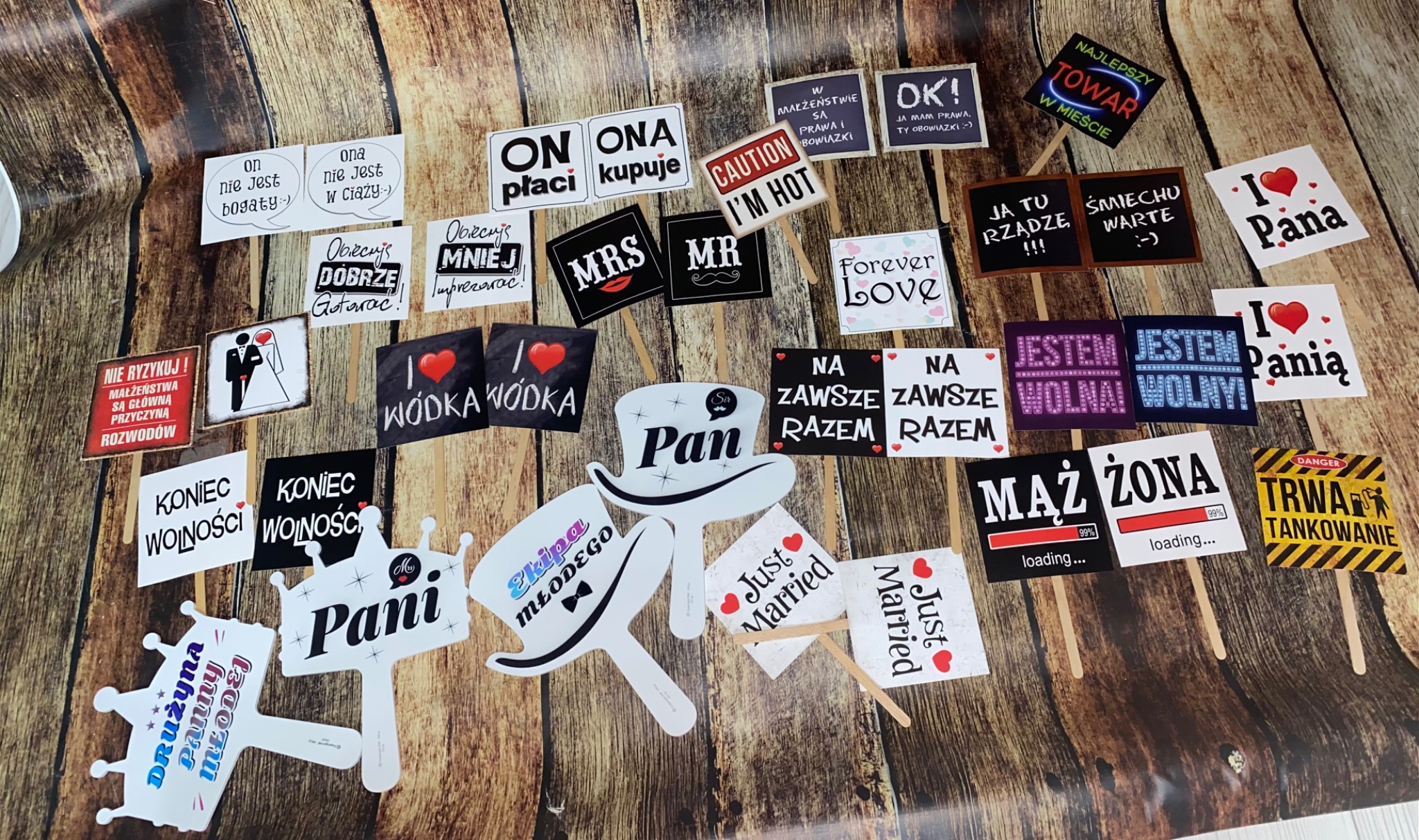 Zestaw rekwizytów do fotobudki na wesele, zawierający różnorodne tabliczki z napisami i grafikami, takie jak 'On płaci, Ona kupuje', 'Mąż 99% loading', 'Żona 99% loading', 'Just Married', 'I love...